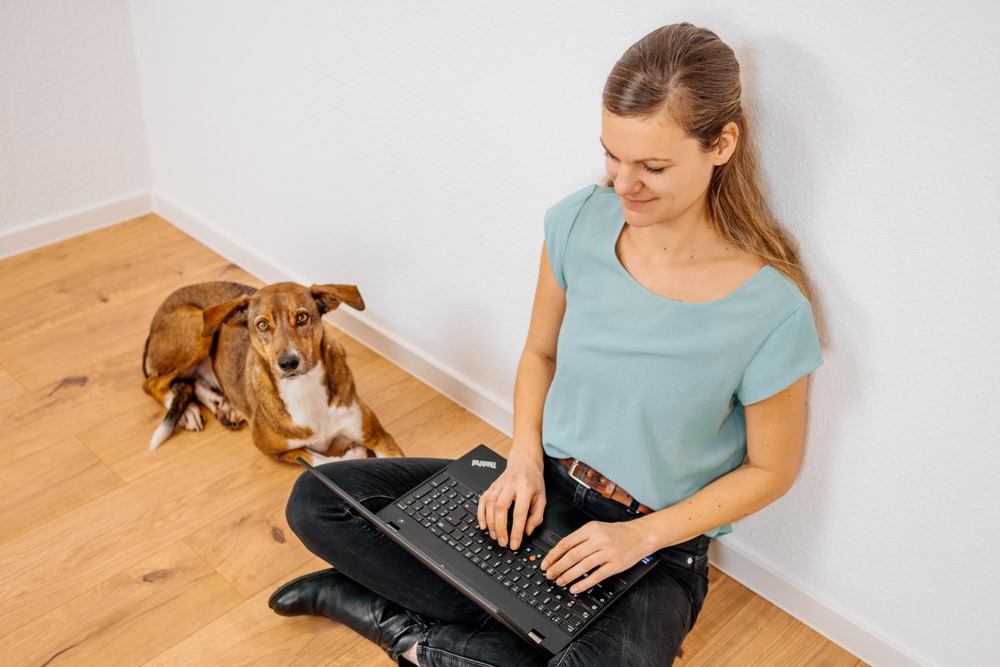 BARF Concept Ernährungsberaterin Teresa sitzt mit Hündin auf Boden, Laptop auf dem Schoß vor einer weißen Wand, Kamerawinkel von schräg oben