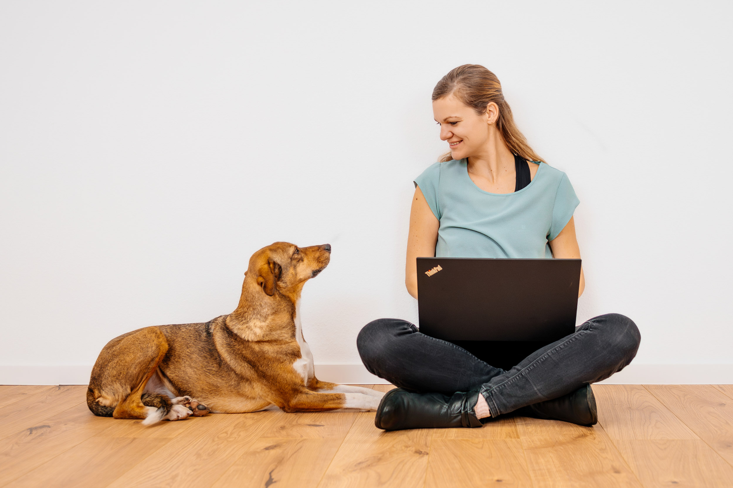 BARF-Hundeernährungsberaterin Teresa sitzt vor weißer Wand auf Boden, Laptop auf Schoß, Hund sitzt neben ihr