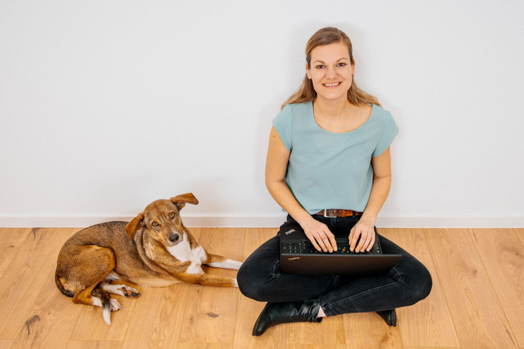 BARF-Hundeernährungsberaterin Teresa sitzt vor weißer Wand auf Boden, Laptop auf Schoß, Hund sitzt neben ihr