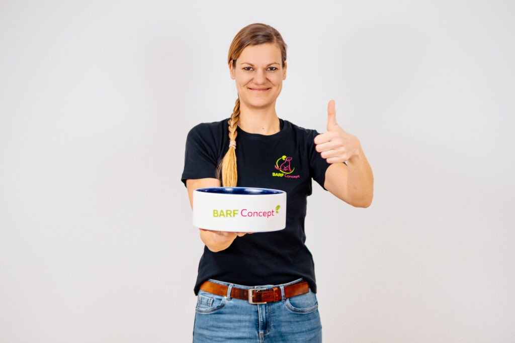 BARF-Concept Inhaberin Teresa trägt schwarzes T-Shirt, geflochtene Haare und hält Hundenapf mit BARF-Futter in der Hand, Daumen hoch